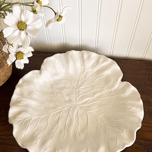 May include: A white ceramic leaf-shaped serving platter with detailed vein patterns. The platter is on a dark wooden surface, with white flowers in a wicker basket in the background. A decorative piece for serving food.