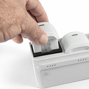 May include: A white charging station with three charging ports. A hand is inserting a small battery into one of the ports. The station has a series of indicator lights and a power button.