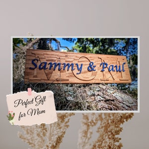 May include: A wooden sign with the words "MOM" carved into it. The sign is painted blue and says "Sammy & Paul" underneath. The sign is sitting on a tree branch.
