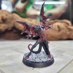 May include: A red and black painted miniature figurine of a demon with wings, standing on a grey stone base.