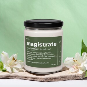 Puede incluir: Una vela blanca en un tarro de cristal con tapa negra. La etiqueta dice "magistrate" con una definición y el nombre "Jennifer". La vela es de 255 g y está sobre una losa de piedra con flores blancas.