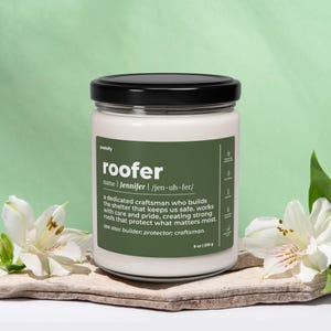 Könnte beinhalten: Eine weiße Kerze in einem Glas mit schwarzem Deckel. Das grüne Etikett trägt den weißen Text "roofer" und eine Beschreibung des Berufs. Die Kerze ist 255 g. Weiße Blumen und eine Holzplatte sind im Vordergrund.