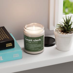 May include: A white candle with a green label that reads "court clerk" with the definition "The heartbeat of the courtroom. Jennifer/jen-uh-fet/ Orchestrating and supporting the wheels of justice, ensures every detail is attended to with care and precision, making the legal process smoother for all.  Case organizer, helper, advocate."