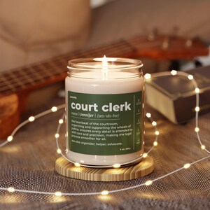 May include: A white candle with a green label that reads "court clerk" with the definition "the heartbeat of the courtroom, organizing and supporting the wheels of justice, ensures every detail is attended to with care and precision, making the legal process smoother for all. See also: organizer, helper, advocate." The candle is lit and sitting on a wooden coaster.