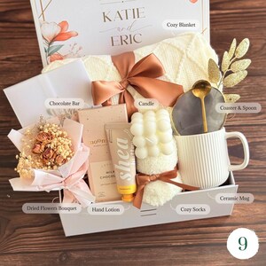 May include: A gift box containing a cream-colored knit blanket with a copper ribbon, a dried flower bouquet, a milk chocolate bar, a shea butter hand lotion, a white bubble candle, a pair of fuzzy socks, and a white ribbed mug with a gold spoon.