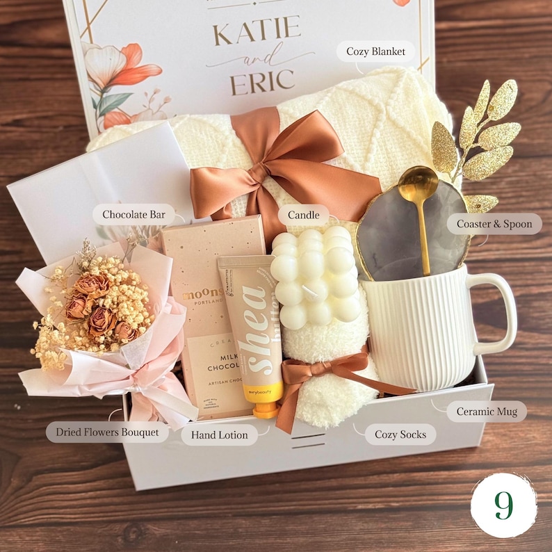 May include: A gift box containing a cream-colored knit blanket with a copper ribbon, a dried flower bouquet, a milk chocolate bar, a shea butter hand lotion, a white bubble candle, a pair of fuzzy socks, and a white ribbed mug with a gold spoon.