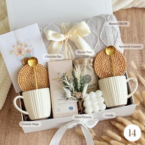 May include: A gift box with a cream-colored blanket (50x70 inches), two ceramic mugs, gold spoons, coasters, a chocolate bar, dried flowers, a candle, and a tea bag. The box is tied with a white ribbon.