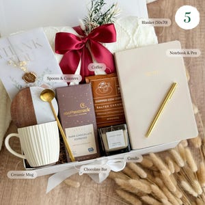 May include: A white gift box filled with a variety of items, including a ceramic mug, a chocolate bar, a candle, coffee, a notebook and pen, and a blanket. The box is decorated with a red ribbon and a wooden coaster.