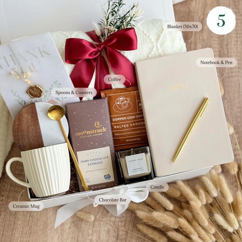 May include: A white gift box filled with a variety of items, including a ceramic mug, a chocolate bar, a candle, coffee, a notebook and pen, and a blanket. The box is decorated with a red ribbon and a wooden coaster.