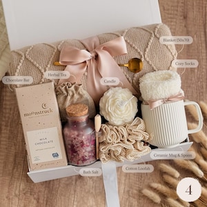 May include: A gift box containing a beige textured blanket measuring 50x70 inches, a Moonstruck milk chocolate bar, a small burlap bag of herbal tea, a glass jar of bath salt with a cork lid, a flower-shaped candle, a cotton loofah, and a white ribbed ceramic mug with a spoon and fuzzy socks.
