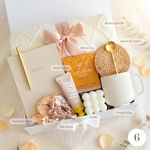 May include: A gift box containing self-care products. It features a cream blanket (127x178 cm), a notebook and pen, herbal tea, hand lotion, a candle, hair bands, a ceramic mug, and a spoon with a coaster. The box is tied with a bow.