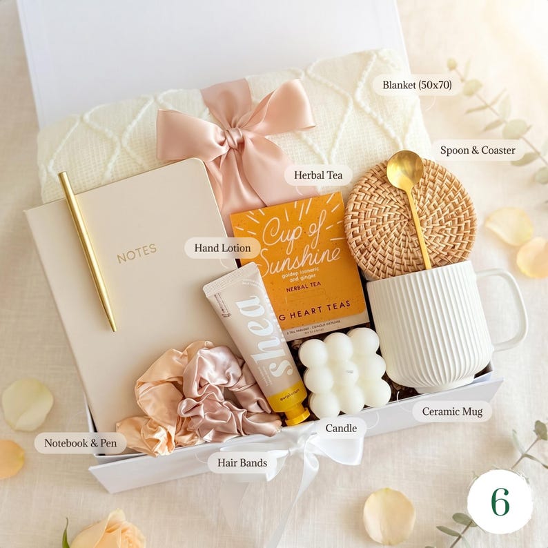 May include: A gift box containing self-care products. It features a cream blanket (127x178 cm), a notebook and pen, herbal tea, hand lotion, a candle, hair bands, a ceramic mug, and a spoon with a coaster. The box is tied with a bow.
