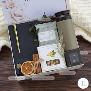 May include: A gift box with a notebook, gold pen, calming tea, roasted pistachios, a tumbler, a candle, dried orange slices, and cinnamon sticks. The box has floral designs and is on a wooden surface.