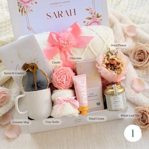 May include: A gift box filled with a variety of items, including a ceramic mug, cozy socks, a candle, chocolate, hand cream, and whole leaf tea. The box is decorated with a pink bow and a card that says "Congratulations Sarah."