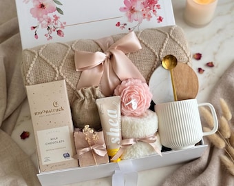 May include: A gift box with a floral design and the name "SARAH" on the lid. Inside, a beige blanket, a mug, a candle, chocolate, soap, and other self-care items are arranged. A pink bow and ribbon add a decorative touch.