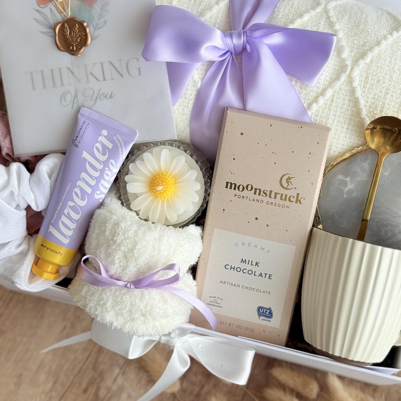 May include: A gift box containing self-care products. It features lavender hand cream, a daisy candle, a milk chocolate bar, a white mug with a gold spoon, and a card reading "Thinking Of You". The box is adorned with white and lilac ribbons and a soft white teddy bear.