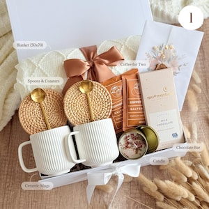 Peut inclure: Coffret cadeau avec une couverture blanche (127x178 cm), deux mugs en c&eacute;ramique blancs, deux cuill&egrave;res dor&eacute;es, deux dessous de verre, des sachets de caf&eacute;, une barre chocolat&eacute;e et une bougie. Le coffret est nou&eacute; avec un ruban.
