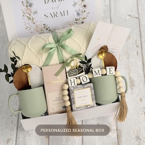May include: A gift box containing a cream cable-knit blanket tied with a green ribbon, two sage green ribbed mugs with gold spoons, a Moonstruck milk chocolate bar, a box of Earl Grey tea, a wooden coaster, and a wooden bead garland with HOME blocks.