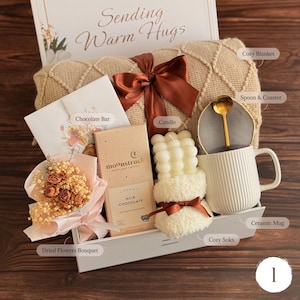 May include: A gift set containing a beige textured blanket with a brown ribbon, a Moonstruck milk chocolate bar, a bubble candle, a white ribbed ceramic mug with a gold spoon and gray coaster, a dried flower bouquet, and a card reading Sending Warm Hugs.