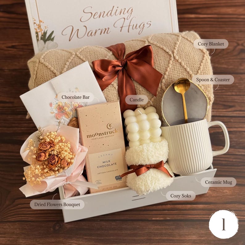 May include: A gift set containing a beige textured blanket with a brown ribbon, a Moonstruck milk chocolate bar, a bubble candle, a white ribbed ceramic mug with a gold spoon and gray coaster, a dried flower bouquet, and a card reading Sending Warm Hugs.