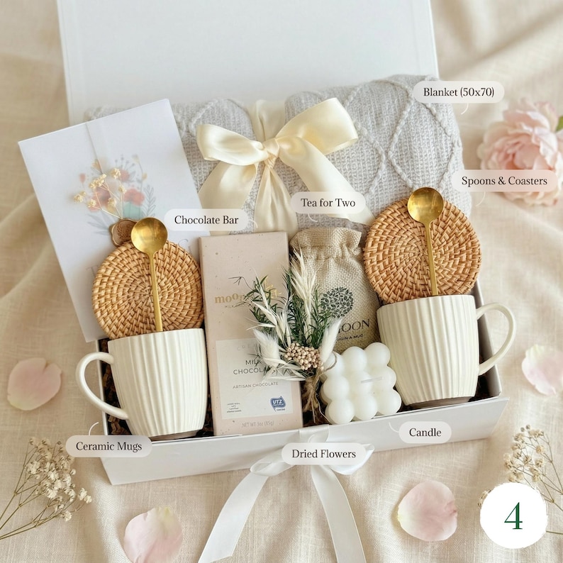 May include: A gift box with a white blanket, two ceramic mugs, coasters, gold spoons, a chocolate bar, dried flowers, a candle, and a card. The blanket measures approximately 19.7 x 27.6 inches. The box is tied with a cream ribbon.
