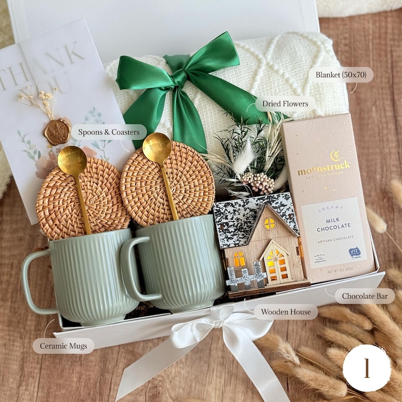 May include: A gift box with a white bow, containing a cream-colored blanket (50x70 inches), two green ceramic mugs, two woven coasters with gold spoons, a chocolate bar, and a wooden house. The box also includes a card that says "THANK YOU".