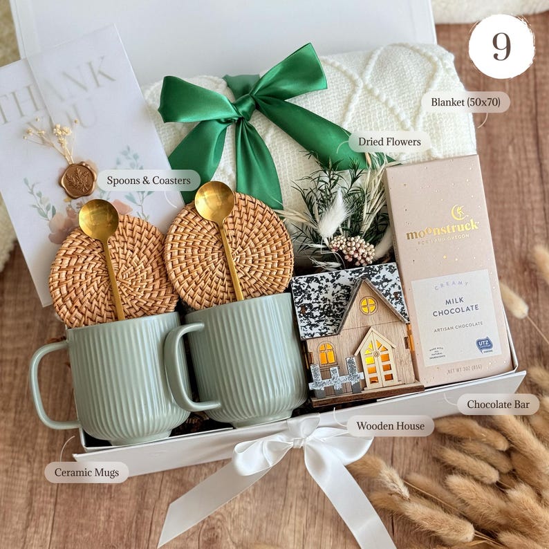 May include: A gift box containing two ribbed sage green ceramic mugs, two round woven rattan coasters, two gold-toned spoons, a white knit blanket with a green ribbon, a small wooden house, dried flowers, and a Moonstruck milk chocolate bar.