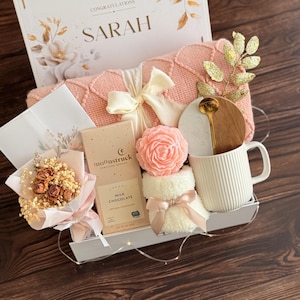 May include: A gift basket with a pink knitted blanket, a white mug, a bar of milk chocolate, a small bouquet of dried flowers, and a rose-shaped soap. The basket also includes a decorative gold spoon and a card that says "Congratulations Sarah".