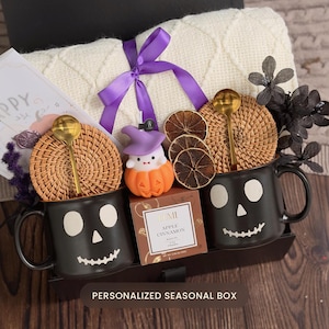 May include: A gift box containing two black skull-faced mugs with gold spoons and woven rattan lids, a cream-colored knit blanket tied with a purple ribbon, a ghost-in-a-pumpkin figurine, dried orange slices, and a box of Apple Cinnamon tea.