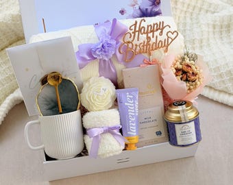 May include: A gift box for Sarah containing a white ribbed mug with a gold spoon and dark coaster, a white rose candle, a small white towel with a purple ribbon, a lavender hand cream, a milk chocolate bar, Earl of Lavender tea, and dried flowers.