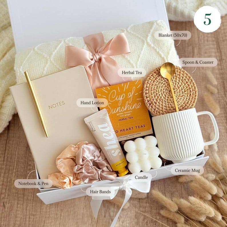 May include: A white gift box filled with self-care items, including a white ceramic mug, a candle, a notebook and pen, a blanket, hand lotion, hair ties, and herbal tea. The box is decorated with a pink ribbon and the text "Cup of Sunshine" on a yellow card.