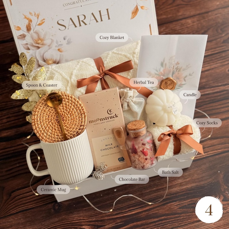 May include: A gift box containing a white ribbed ceramic mug, a rattan coaster with a gold spoon, a Moonstruck milk chocolate bar, a glass jar of bath salts with dried petals, a white pumpkin-shaped candle, cozy socks, and a cream knit blanket.