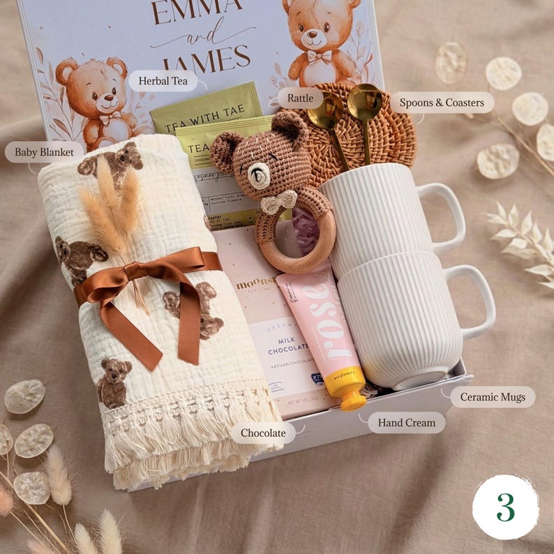 May include: A gift box with baby-themed items. Includes a baby blanket with teddy bear prints, a rattle, two white ceramic mugs, chocolate, hand cream, tea, and spoons. The box is decorated with teddy bear illustrations and the names "Emma and James".