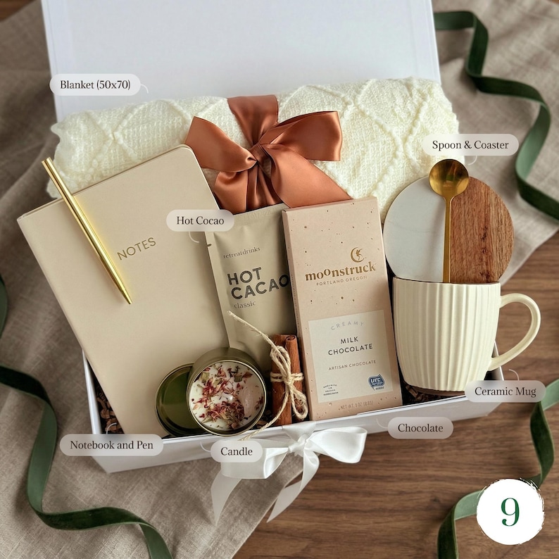 May include: A gift box with a cream-colored blanket (50x70 inches), notebook, pen, hot cocoa, chocolate bar, candle, mug, spoon, and coaster. The box is tied with a ribbon and bow, perfect for a cozy gift.