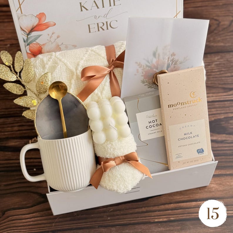 May include: A gift basket filled with a white mug, gold spoon, candle, blanket, hot cocoa, and chocolate bar. The basket is decorated with a ribbon and a card that says "Katie and Eric".