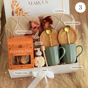 May include: A gift box containing a beige textured blanket with a brown ribbon, two sage green ribbed ceramic mugs with gold spoons, two round woven coasters, a box of Rustic Bakery pumpkin cookies, a jar of honey, a honey dipper, and a pumpkin candle.