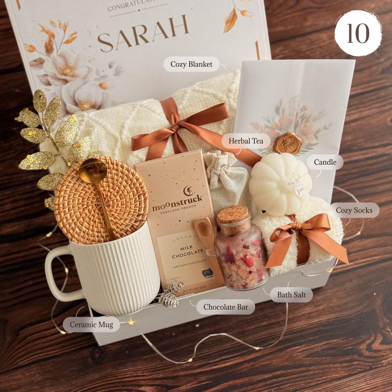 May include: A gift box containing a white ribbed ceramic mug with a rattan coaster and gold spoon, a Moonstruck milk chocolate bar, a glass jar of bath salts, a white pumpkin-shaped candle, a cream-colored knit blanket, and herbal tea.
