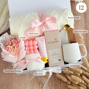 May include: A gift box filled with a cream-colored blanket, pink candle, hand lotion, chocolate bar, dried flower bouquet, cozy socks, and a ceramic mug. A wooden spoon and coaster are also included. The blanket measures 50x70 inches.