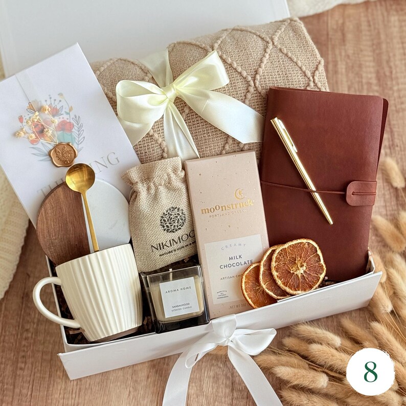 May include: A gift box filled with a variety of items. Includes a cream-colored mug, a small candle, a chocolate bar, a journal, a pen, a small bag, a greeting card, a wooden coaster, a gold spoon, and dried orange slices. The box is tied with a white ribbon.