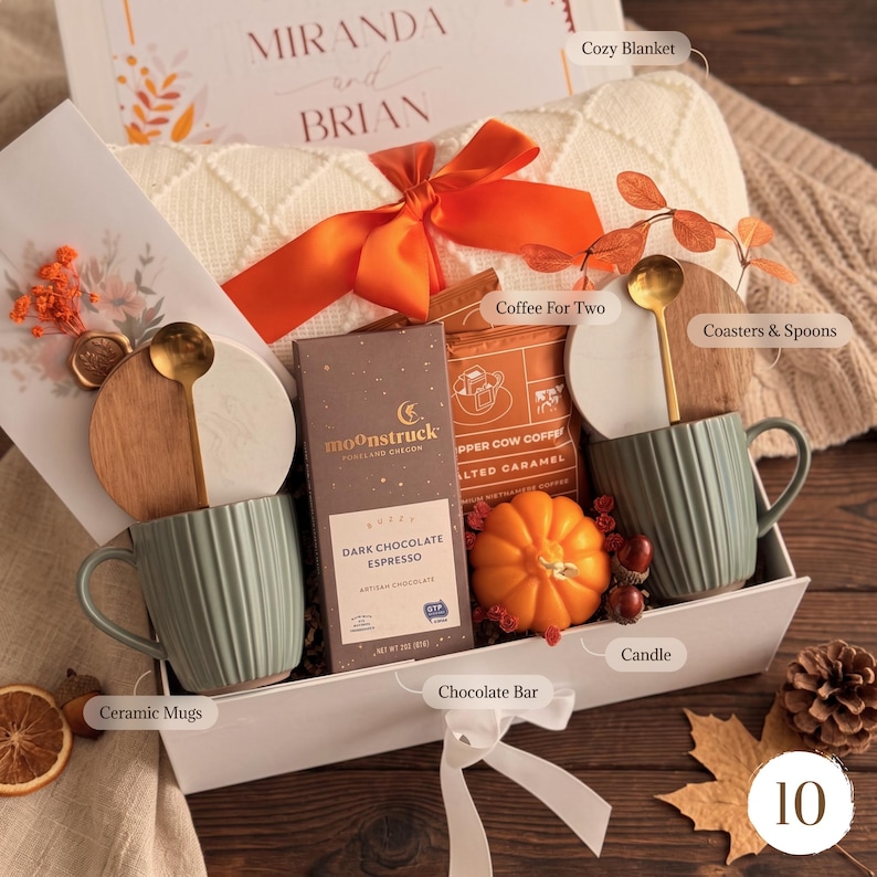 May include: A gift box containing two sage green ribbed ceramic mugs, two round wooden coasters with gold spoons, a Moonstruck dark chocolate espresso bar, a bag of Copper Cow salted caramel coffee, and a pumpkin-shaped candle with a knit blanket.