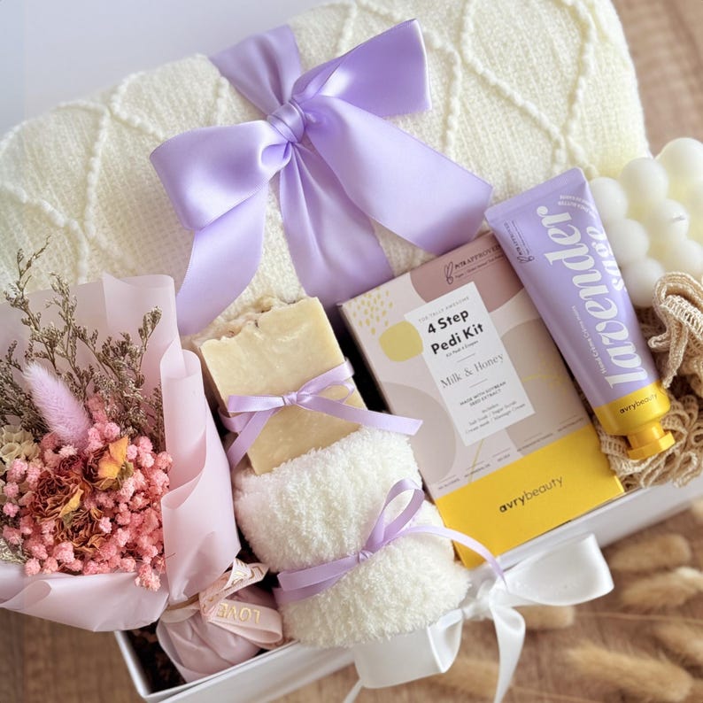 May include: A gift box containing self-care products. It features a white blanket with a lilac bow, a dried flower bouquet, a bar of soap, a foot scrub, a lavender hand cream tube, and a 4-step pedi kit.