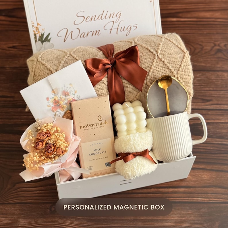 May include: A gift box containing a beige textured throw blanket with a rust-colored ribbon, a Moonstruck milk chocolate bar, a white ribbed mug with a gold spoon, a bubble candle, a small dried flower bouquet, and a card reading Sending Warm Hugs.
