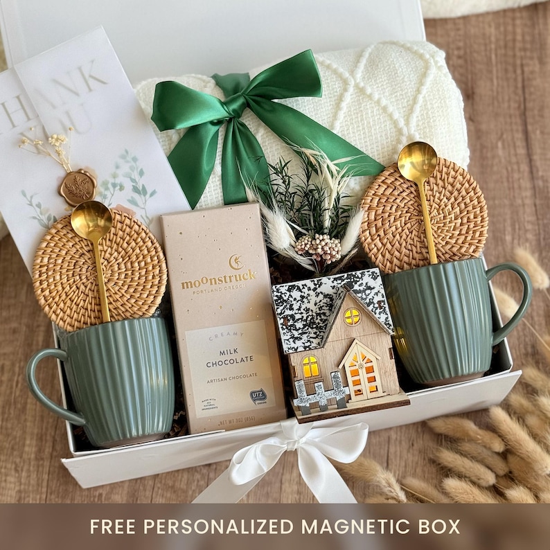 May include: A gift box with a white lid, containing two green mugs with gold spoons, woven coasters, a chocolate bar, a small wooden house, and a cream-colored blanket. The box is tied with a green ribbon and a white bow. The text "THANK YOU" is visible.