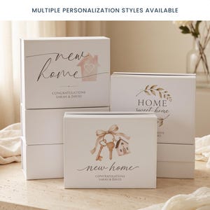 May include: White rectangular gift boxes with hinged lids featuring various designs. One box shows a watercolor house and the text "new home CONGRATULATIONS SARAH & DAVID". Another displays a key and house illustration with "new home".