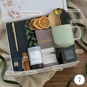 May include: A gift box filled with a variety of items. It includes a green ceramic mug, a notebook with a gold pen, herbal tea, dark chocolate, a candle, dried orange slices, a spoon, and a coaster. The box is decorated with floral accents.