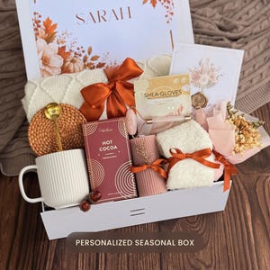 May include: A gift box containing a white ribbed mug with a gold spoon, a box of hot cocoa, a pair of shea butter gloves, a pink jade roller, a ribbed candle, a white fuzzy pair of socks, a small dried flower bouquet, and a white knit blanket.