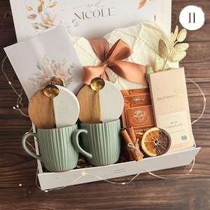 May include: A gift box containing two sage green ribbed mugs with wooden and white lids, two gold spoons, a white knit blanket with a copper ribbon, a Moonstruck milk chocolate bar, two coffee packets, cinnamon sticks, and a dried orange slice.