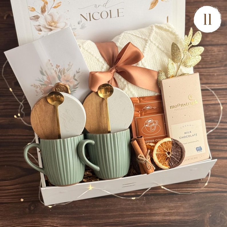 May include: A gift box containing two sage green ribbed mugs with wooden and white lids, two gold spoons, a white knit blanket with a copper ribbon, a Moonstruck milk chocolate bar, two coffee packets, cinnamon sticks, and a dried orange slice.