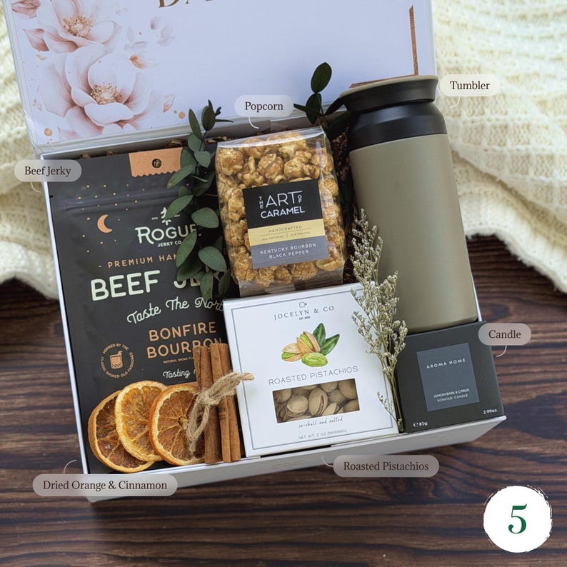May include: A gift box filled with gourmet treats. Includes beef jerky, caramel popcorn, a tumbler, roasted pistachios, a candle, dried orange slices, and cinnamon sticks. The box is white with a floral design.