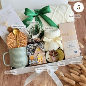 May include: A gift box containing a white knit blanket with a green ribbon, a light green ceramic mug, a rattan coaster, a gold spoon, dried flowers, white cozy socks, a wooden house ornament, a candle, and a Moonstruck milk chocolate bar.